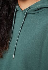 Zelená mikina s kapucí vyrobená z měkkého materiálu, s tkaničkami se stříbrnými konci a designem klokaní kapsy. Hladká textura, volný střih.