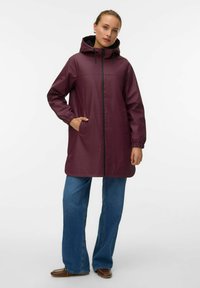 Parka impermeabile color bordeaux con cappuccio, cordoncini regolabili, due tasche laterali e chiusura con zip, abbinata a jeans a gamba larga blu.