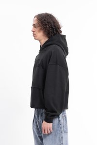 Jeune personne avec des cheveux bouclés portant un hoodie noir surdimensionné et un jean bleu clair, se tenant de profil sur un fond blanc.
