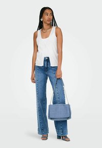 Top bianco senza maniche con un dettaglio annodato sul davanti, abbinato a jeans blu a vita alta. Il modello tiene una borsa blu con texture. Sandali blu con cinturini.