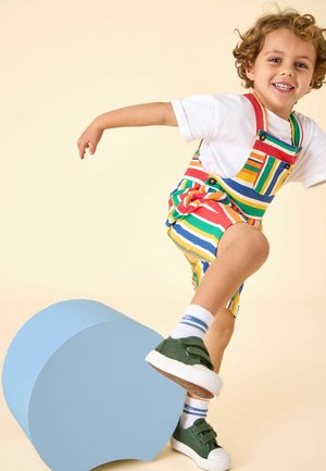 Noor laps lokkis juustega, kandes triibulisi pükse ja rohelisi tossusid, tasakaalu hoides ühe jalaga suurel sinisel silindrilisel objektil.