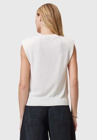Witte mouwloze sweater met geribde zoom en schouderdetails. Zachte textuur met subtiele stiksels op de achterkant. Gecombineerd met donkere broek.