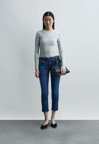 Kvinne iført en grå langermet topp, avkuttede blå jeans, svarte flate sko, holder en liten svart håndveske, stående mot en enkel lys bakgrunn.