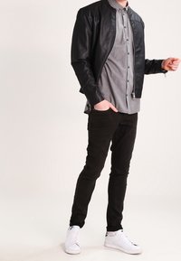 Sort læder bomberjakke over en grå skjorte med knapper, kombineret med sorte slim-fit jeans og hvide sneakers. Glat tekstur, afslappet design.