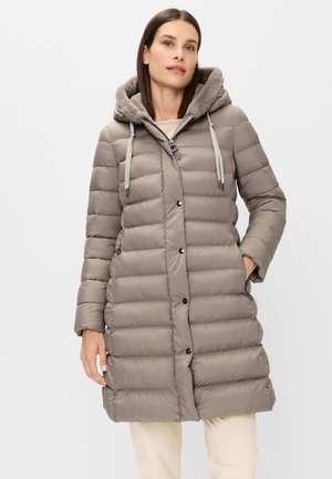 Frau, die eine lange, taupefarbene gesteppte Steppjacke mit Kapuze, die mit Pelz gefüttert ist, vorn mit Knöpfen und seitlichen Reißverschlusstaschen trägt, steht vor weißem Hintergrund.