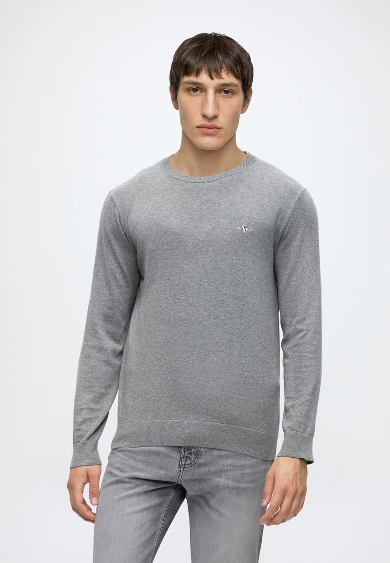 Young man with short brown hair wearing a plain grey long-sleeve sweater and grey jeans, standing against a white background.