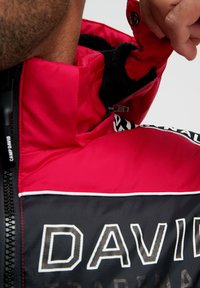 Rote und schwarze gepolsterte Jacke mit hohem Kragen, ausgestattet mit einem Reißverschluss, Fleece-Futter und weißen Logo-Akzenten am Saum.