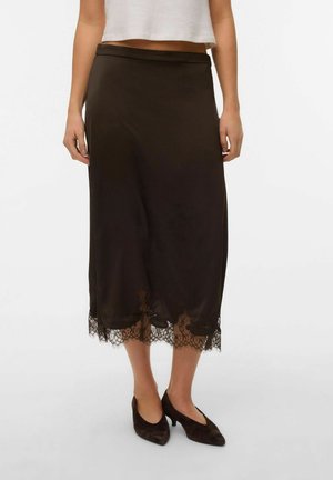 Mujer con falda midi negra con detalle de encaje en el dobladillo y zapatos negros de tacón bajo, de pie frente a un fondo liso.