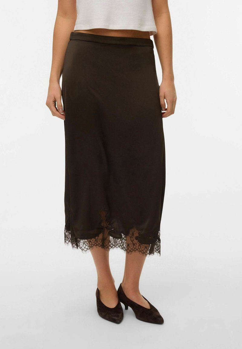 Femme portant une jupe midi noire avec un détail en dentelle à l'ourlet et des chaussures noires à talon bas, debout devant un fond uni.