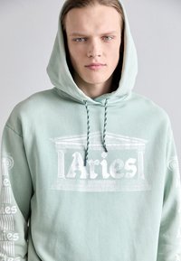 Sweat à capuche vert clair avec des graphiques imprimés blancs, y compris le logo "Aries" et un design architectural. Dispose de cordons de serrage ajustables avec des accents foncés.