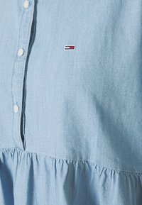 Błękitna bluzka chambray z guzikiem z przodu, marszczoną dolną częścią i małym haftowanym logo w czerwonym, białym i niebieskim kolorze.