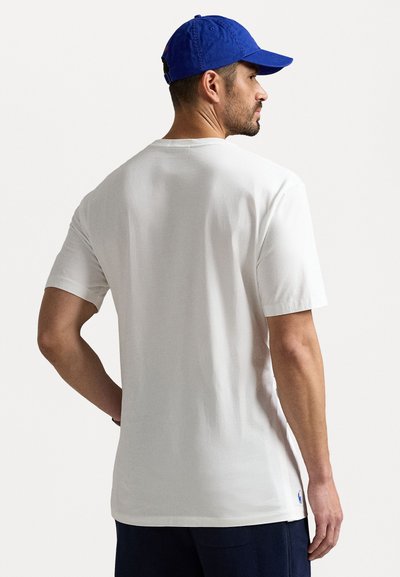 T-shirt blanc à manches courtes avec col rond, en coton, avec des fentes latérales à l'ourlet, porté avec une casquette bleue. Vue arrière.