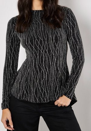 Kobieta ubrana w dopasowaną czarną bluzkę z długim rękawem z srebrnymi falistymi pionowymi paskami, w zestawieniu z czarnymi spodniami, pozująca z jedną ręką na biodrze.