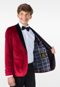 Czerwony aksamitny blazer z czarnymi satynowymi klapami, z pojedynczym guzikiem z przodu i kieszenią. W środku znajduje się wyściółka w czarno-szarym kratkę z humorystycznym przesłaniem.