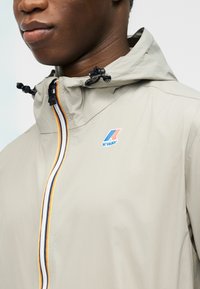 Κοντινό πλάνο ενός ατόμου που φοράει ένα μπεζ μπουφάν K-Way με πολύχρωμο φερμουάρ με ρίγες και ρυθμιζόμενα κορδόνια κουκούλας.