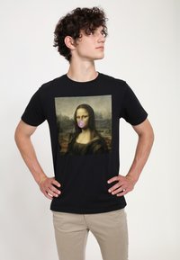 Černé bavlněné tričko s grafikou Mona Lisy, která má na ústech růžovou žvýkačku. Modelka stojí se založenýma rukama v pase.