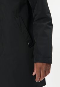 Detail ruky osoby vedle rukávu a kapsy černého kabátu s viditelnou malou textilní značkou.