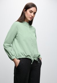 Camicia verde chiaro con colletto rotondo, polsini elasticizzati e orlo arricciato con cordoncino. Abbinata a pantaloni scuri e larghi.