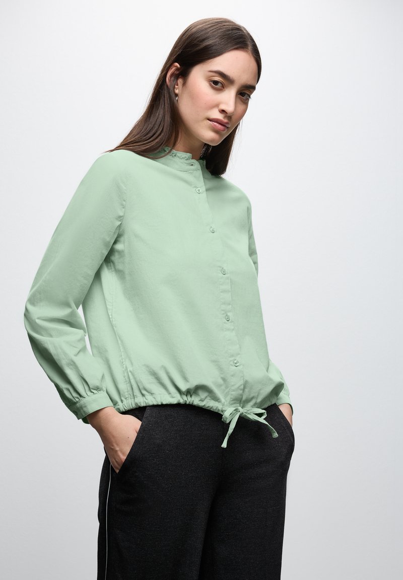 Camicia verde chiaro con colletto rotondo, polsini elasticizzati e orlo arricciato con cordoncino. Abbinata a pantaloni scuri e larghi.