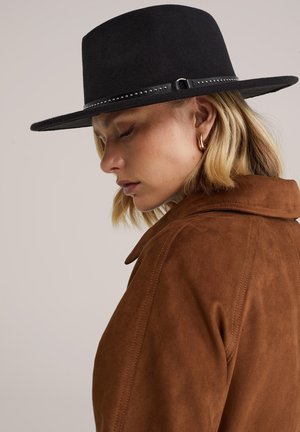 Brūna zamšādas jaka ar apkakli un redzamām šuvēm, kopā ar melnu filca cepuri ar platu malu un kniedētu melnu joslu.