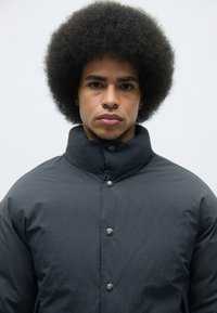 Black puffer jacket with a high collar, featuring silver snap buttons. Smooth, lightweight fabric with a matte finish.