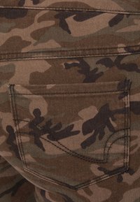 Gros plan sur un tissu camouflage marron et vert avec une poche arrière cousue présentant un design incurvé à double surpiqûre.