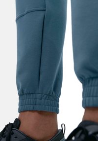 Pantaloni jogger blu con polsini elasticizzati e dettagli delle tasche laterali, realizzati in tessuto morbido. Completati con accenti di scarpe da ginnastica nere ai piedi.