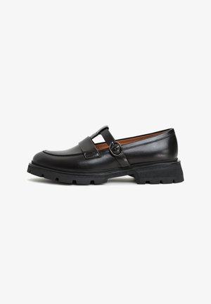Black leather shoe with a round toe, raised front seam, and a wide strap featuring a circular buckle. Chunky rubber sole with treads.