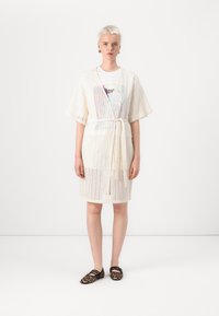 Rochie din dantelă crem cu talie legată, având un model geometric și mâneci scurte, purtată peste un tricou grafic și asociată cu pantofi cu imprimeu leopard.