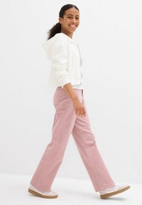 Felpa bianca corta, pantaloni a gamba larga rosa chiaro e sneakers bianche con suole marroni. Sfondo semplice, modella che cammina e sorride.