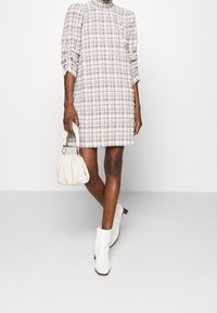 Brown and white checked dress with a high neckline and gathered sleeves, paired with a cream handbag and white ankle boots.