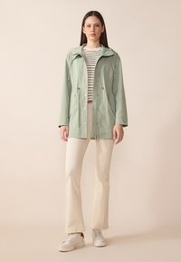 Jachetă waterproof de culoare verde deschis, cu glugă, închidere cu fermoar și șnururi ajustabile. Asortată cu un top cu dungi și pantaloni evazați de culoare crem.