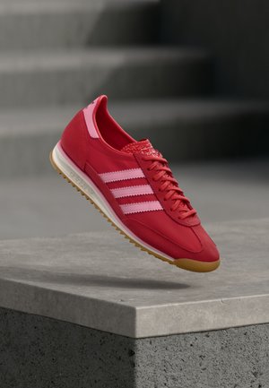 Zapatilla deportiva roja con detalles rosas, materiales de ante y sintéticos, que presenta tres franjas blancas, puntera texturizada y suela de goma color caramelo.