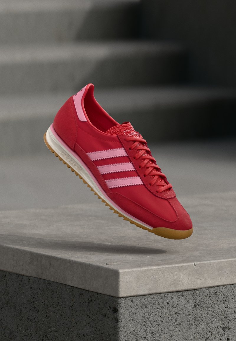 Chaussure de sport rouge avec des accents roses, en daim et matériaux synthétiques, comportant trois bandes blanches, un embout texturé et une semelle en caoutchouc gomme.
