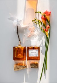 Bouteilles de parfum en verre avec un liquide ambré, des caps blancs, étiquette portant la mention "Sikinos." Entourées de fleurs roses, de cristaux de sel et d'un moulin à vent blanc.
