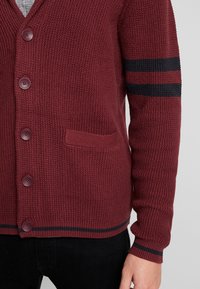 Cardigan bordeaux lavorato a maglia con tessuto testurizzato, caratterizzato da cinque bottoni, una tasca frontale e dettagli a righe nere sulle maniche.