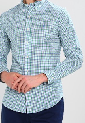 Man draagt een lichtblauw, groen en wit geruit overhemd met lange mouwen en donkerblauwe broek, handen voor zich gevouwen.