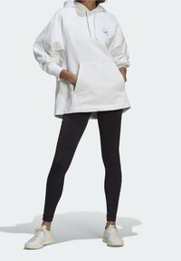 Vit oversized hoodie med en framficka och logotyp, kombinerad med svarta leggings med snörningsdetaljer på sidorna och vita sneakers.