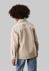 Camicia di corduroy beige con colletto, maniche lunghe e polsini con bottoni, vista dal retro, abbinata a jeans azzurri chiari.