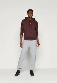 Bordowa bluza z kapturem i kangurzą kieszenią, z białym logo, w połączeniu z jasnoszarymi spodniami dresowymi z bocznymi kieszeniami, wszystkie z miękkiego materiału. Czarne trampki.