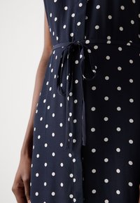 Gros plan d'une robe bleu marine à pois blancs nouée à la taille par une ceinture en tissu assorti.