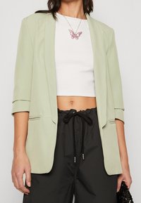 Světle zelené sako s přizpůsobeným střihem a klopovým límcem, nošené přes bílý crop top a doplněné černými kalhotami s šňůrkou v pase. Náhrdelník s přívěskem růžového motýla.