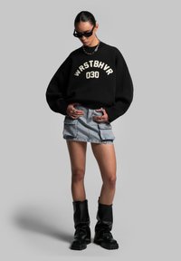 Black knitted sweater with cream logo text, denim cargo mini skirt, and knee-high black boots with textured soles. Accessories include sunglasses.