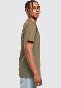 Oliven grøn kortærmet t-shirt i en afslappet pasform, lavet af blødt stof med rund halsudskæring. Bæreren har fletninger og står i profil.