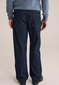 Blugi din denim albastru închis, cu o croială dreaptă, având două buzunare la spate și o textură subtilă. Asortați cu un pulover bleu deschis.