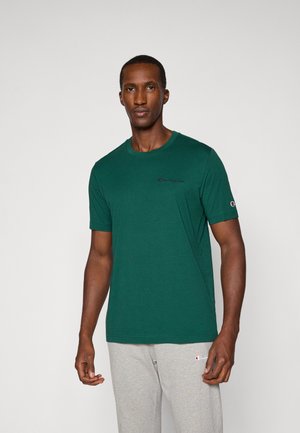 T-shirt en coton vert avec un col rond, une silhouette ajustée et un petit logo en violet sur la poitrine gauche. Porté avec un pantalon de sport gris.