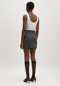 Weißes Tanktop, grauer Nadelstreifen-Minirock, schwarze knielange Socken und schwarze High-Heels, von hinten betrachtet. Glattes Material und figurbetonte Passform.