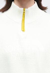 Žena nosící bílý svetr s texturou a čtvrtinovým zipem, žlutým poutkem na zip a jemně vyvýšeným logem na hrudi.