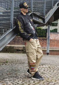 Svart bomberjacka med gul broderi, grafisk t-shirt, beige cargobyxor med tigerpatch och svarta sneakers med vita accenter.