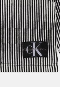 Mustavalkoinen raidallinen kangas, jossa on teksturoitu pinta. Mukana musta etiketti, jossa lukee "Calvin Klein Jeans" ja valkoinen "CK" logo.